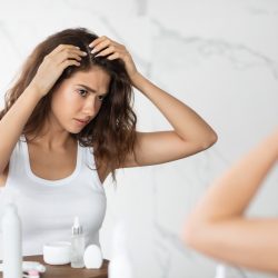 Frustrated Young Lady Searching Hair Flakes Suffering From Dandruff Problem Standing Near Mirror In Bathroom Indoors. Haircare And Head Skin Health Concept. Selective Focus