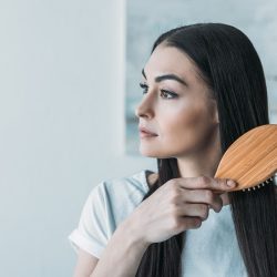 young brunette woman combing hair with hairbrush and looking away